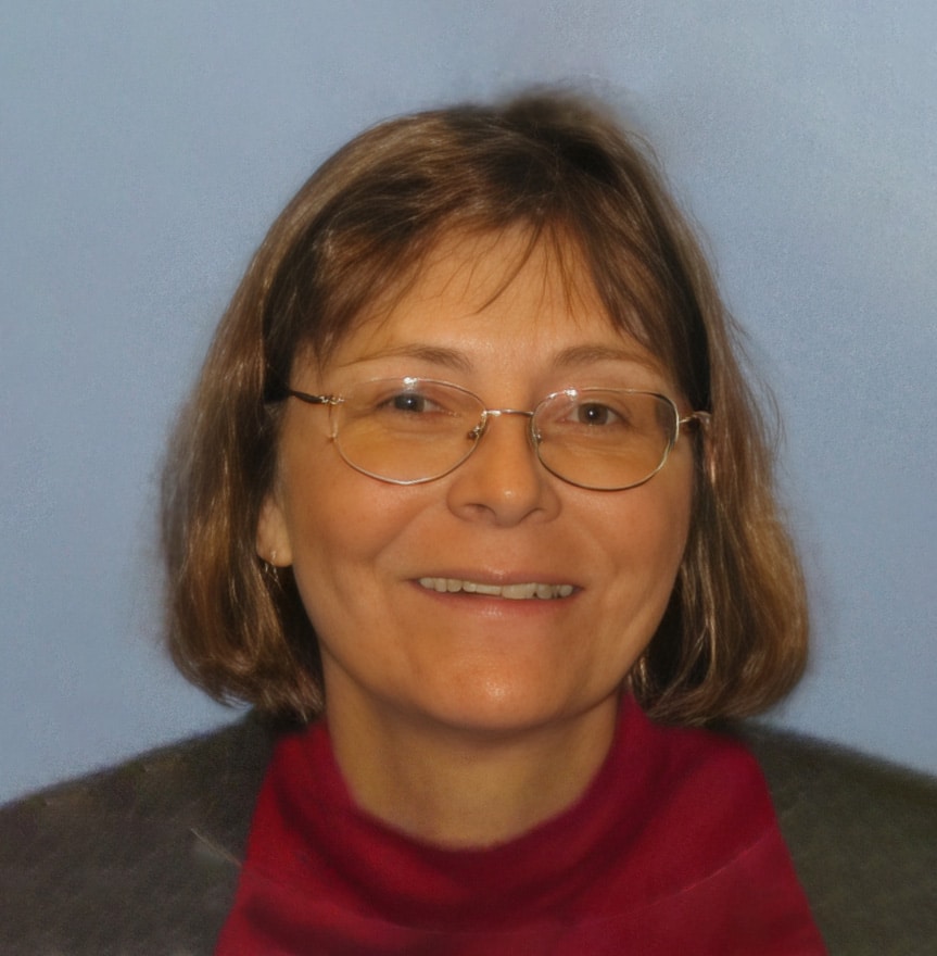 A woman with short brown hair and glasses smiles at the camera. She is wearing a red turtleneck and dark jacket, with a light blue background behind her.