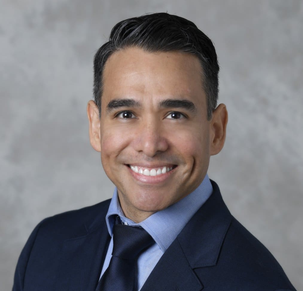 A man with dark hair, wearing a navy blue suit, light blue shirt, and navy tie, smiles in a professional headshot against a light gray, textured background.