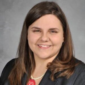 A woman with straight brown hair wearing a dark blazer, colorful blouse, and pearl necklace is smiling in front of a neutral gray background.