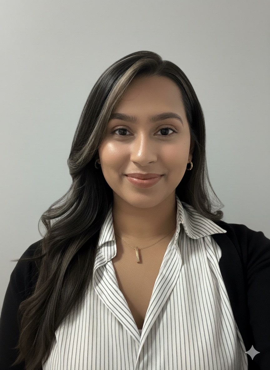 A woman with long, wavy brown hair smiles at the camera. She is wearing a white pinstripe blouse, a black blazer, gold hoop earrings, and a gold necklace, standing in front of a plain gray background.