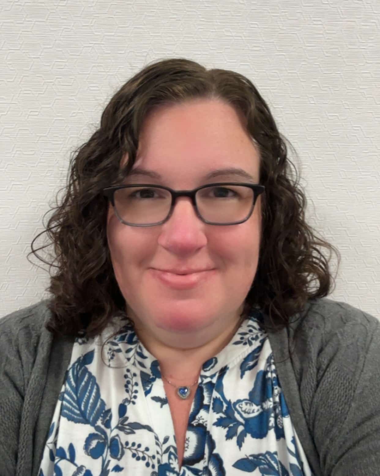 A woman with curly brown hair and glasses smiles at the camera. She is wearing a gray cardigan over a white blouse with blue floral patterns, and a blue pendant necklace. The background is a white textured wall.