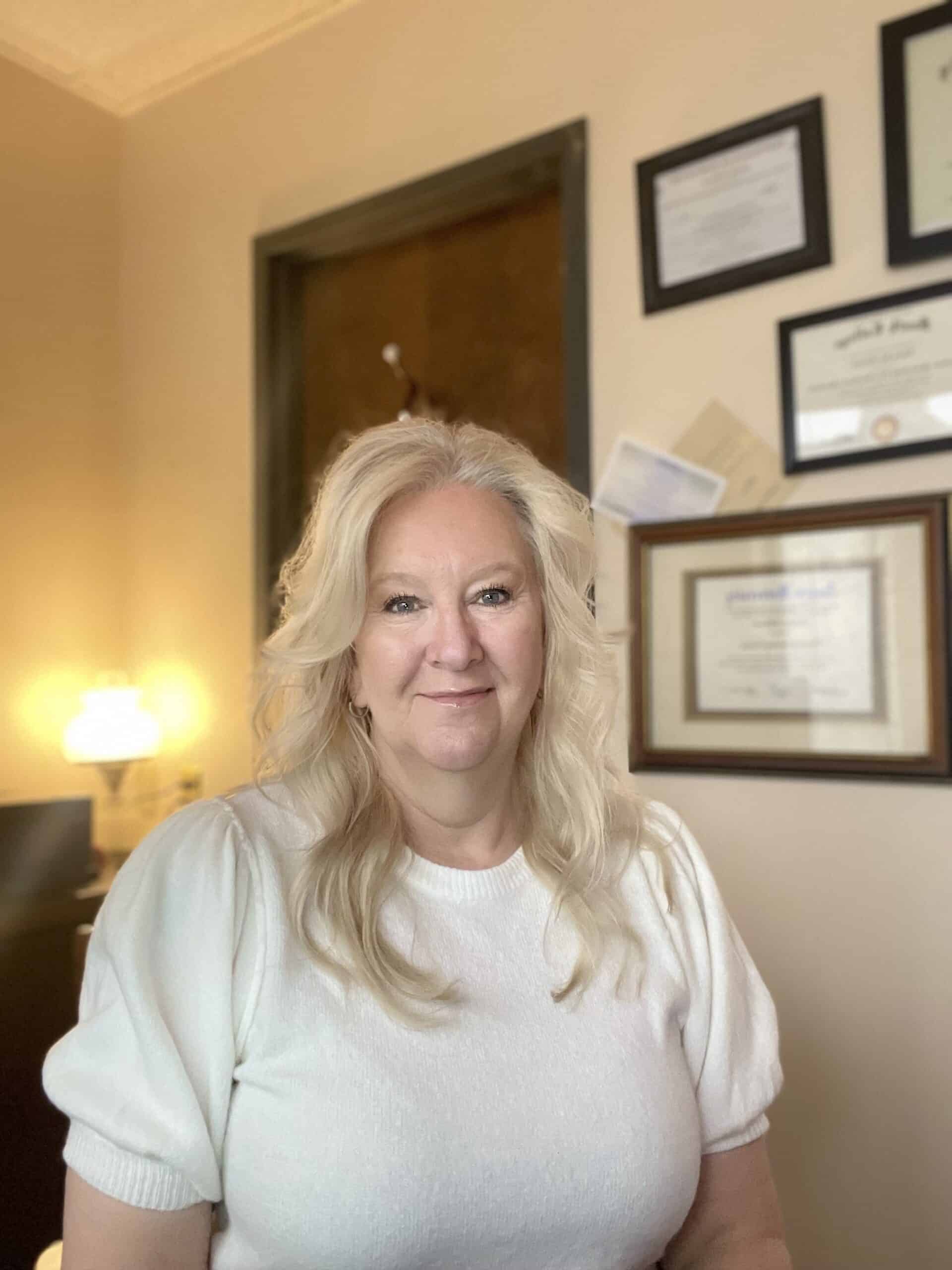 A woman with long blonde hair wearing a white top is smiling in an office. Framed certificates and documents are displayed on the wall behind her.