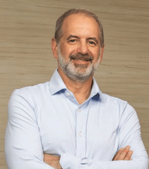A middle-aged man with short gray hair and a beard stands smiling with his arms crossed, wearing a light blue button-up shirt, in front of a beige textured background.