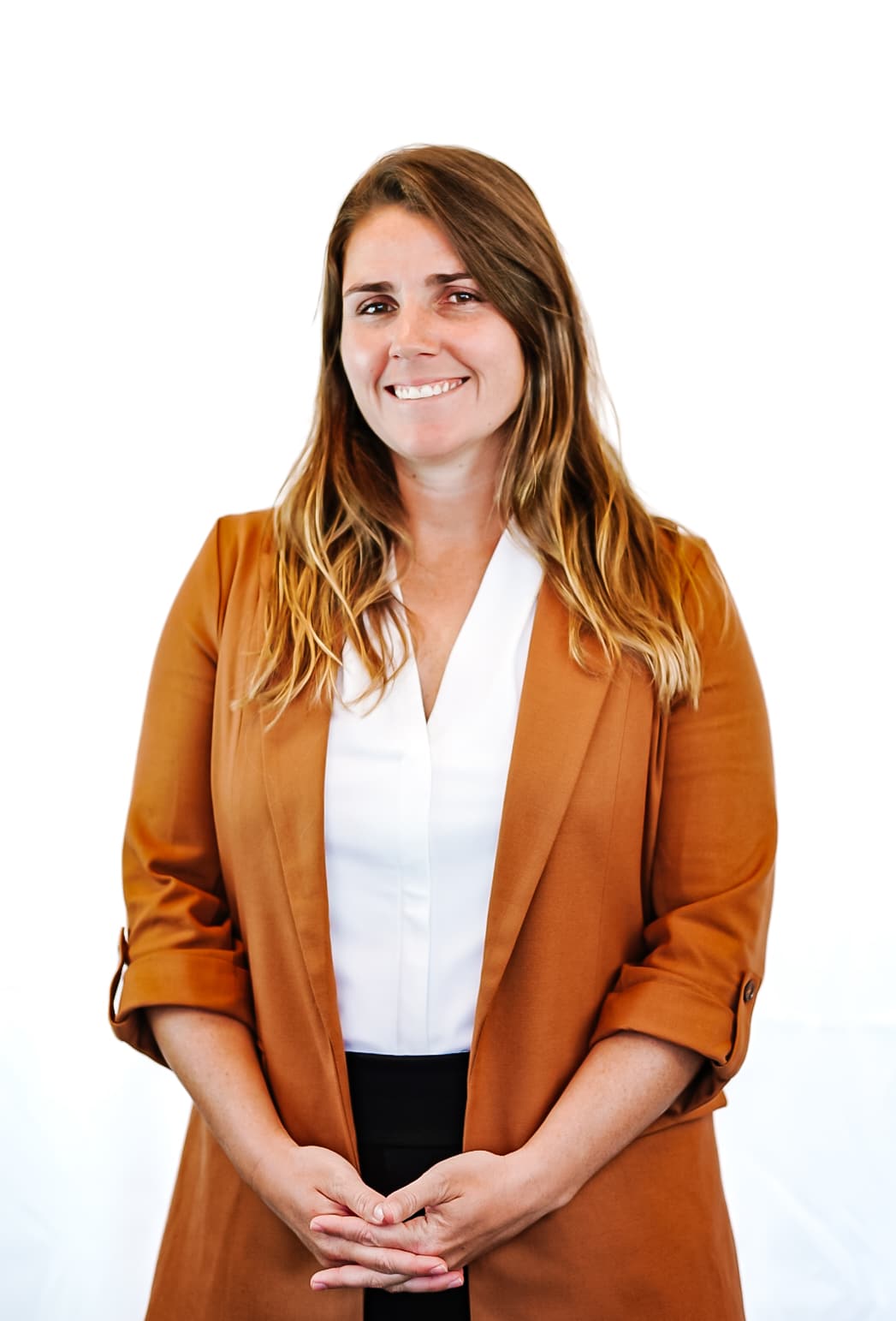 A woman with long brown hair wearing a white blouse and a brown blazer stands smiling with her hands folded in front of her against a plain white background.