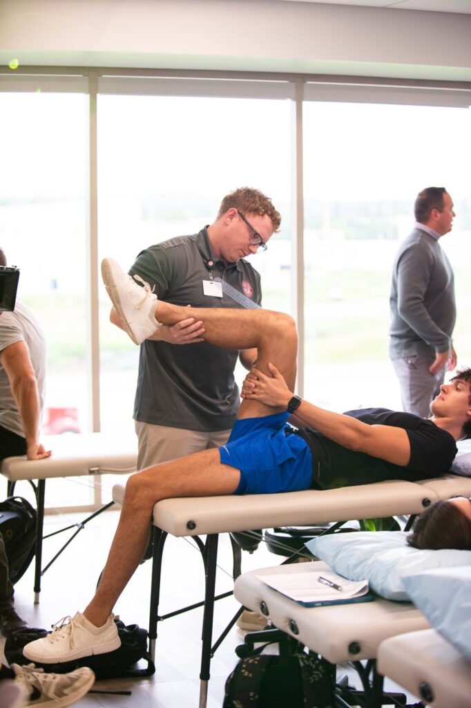 A physical therapist in a gray polo and glasses is assisting a male patient in blue shorts by stretching his leg on a treatment table in a bright clinic room with large windows.