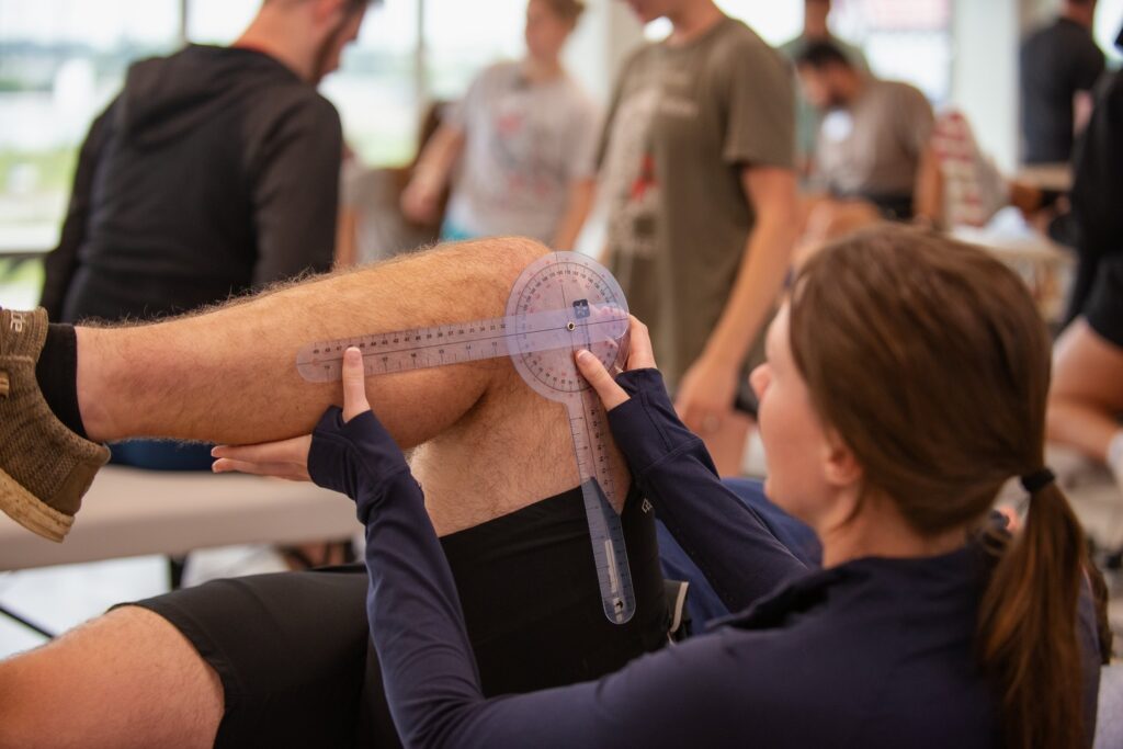 A physical therapist uses a goniometer to measure a patient’s knee angle while the patient lies on a table. Other people are visible in the background, engaged in similar activities.