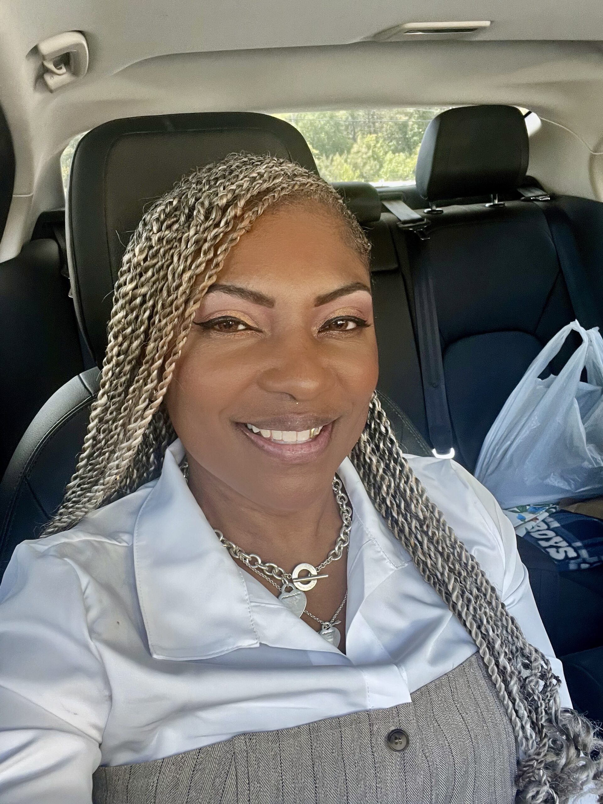 A woman with long braids smiles while sitting in a car. She is wearing a white collared shirt and layered necklaces.