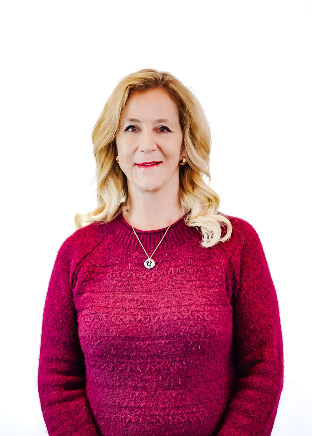 A woman with long blonde hair wearing a maroon sweater and a pendant necklace stands smiling in front of a plain white background.