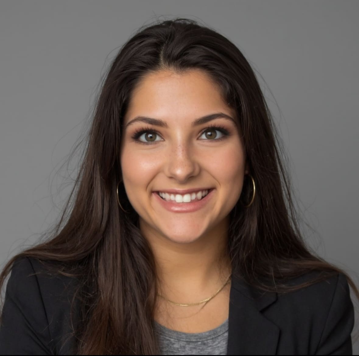A young woman with long brown hair, wearing a black blazer, gray top, hoop earrings, and a gold necklace, smiles at the camera against a plain gray background.