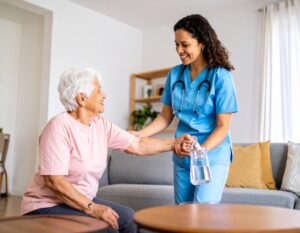 A nurse in blue scrubs smiles while helping an elderly woman, who is seated and smiling back. The nurse holds a bottle of water and supports the woman’s arm in a bright, cozy living room.