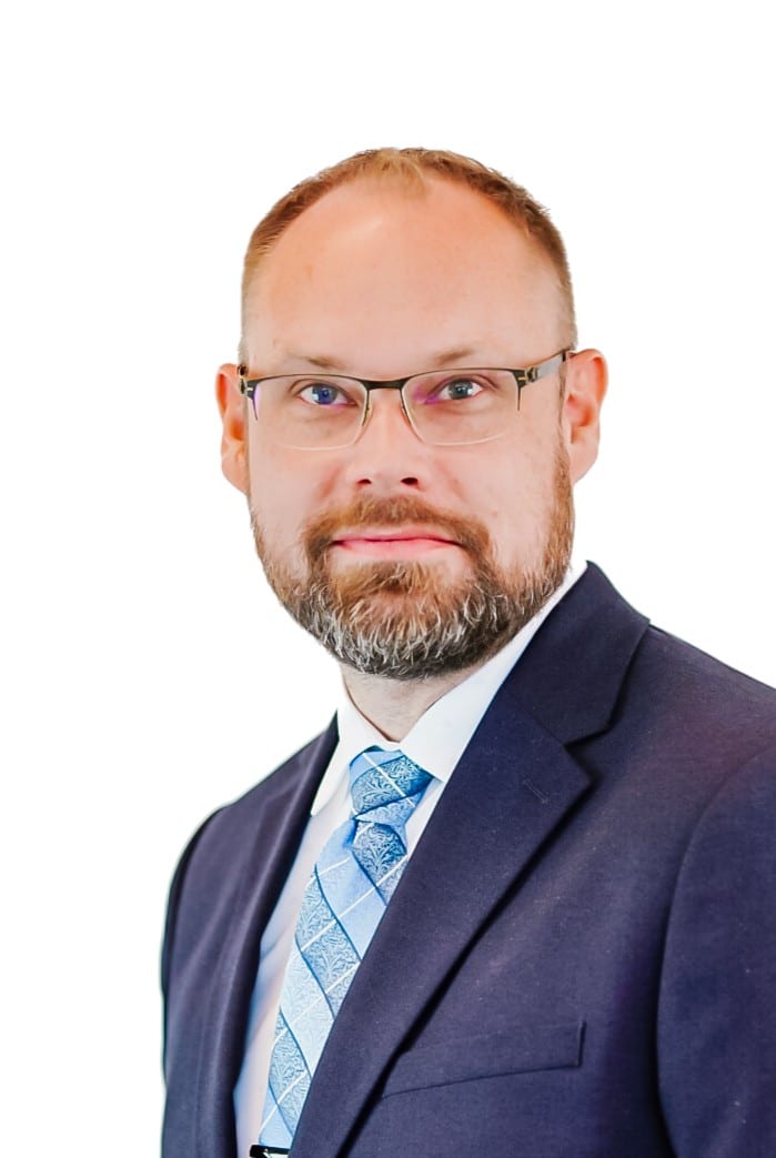 A man with short light brown hair, a beard, and glasses is wearing a dark navy suit, white shirt, and a patterned blue tie. He is looking at the camera against a plain white background.