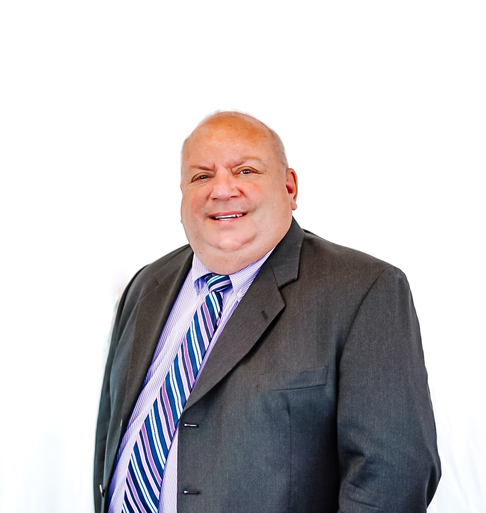 A man wearing a dark suit, striped tie, and light-colored shirt smiles while standing against a plain white background.