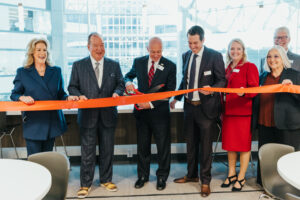 Seven people in business attire smile as a man in the center cuts a red ribbon with large scissors, indicating a ribbon-cutting ceremony in a modern indoor setting.