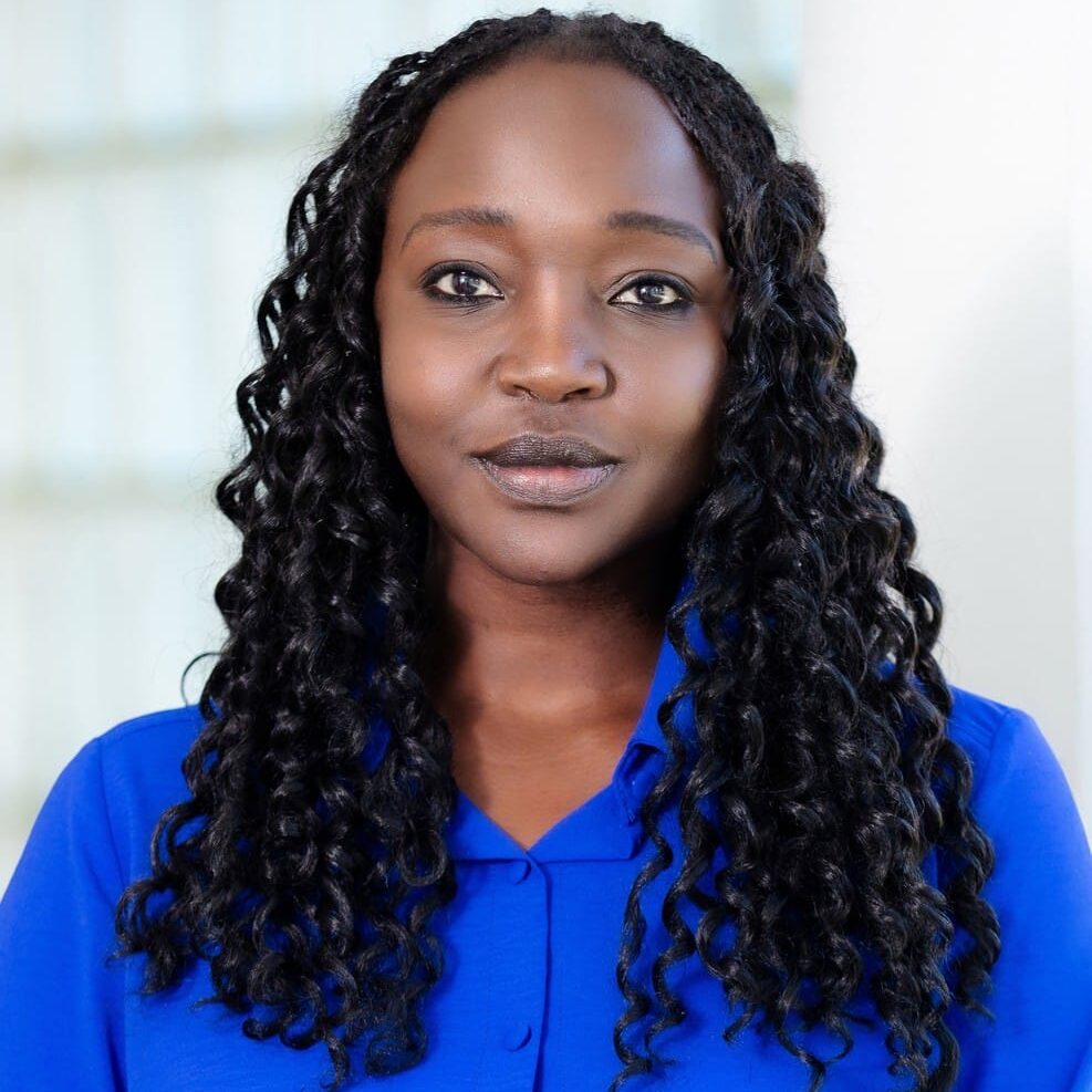 A woman with long curly hair wearing a bright blue button-up shirt stands indoors in front of a light-colored background, looking directly at the camera.