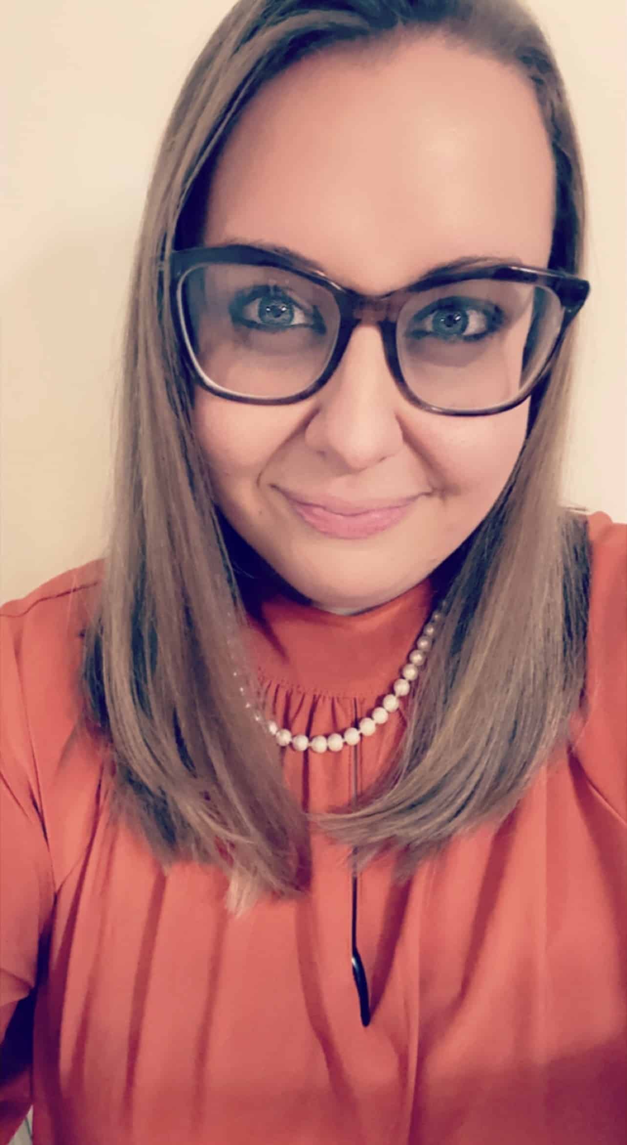 A woman with straight, shoulder-length light brown hair smiles at the camera. She is wearing glasses, a pearl necklace, and an orange blouse with a high, gathered neckline. The background is plain and light-colored.