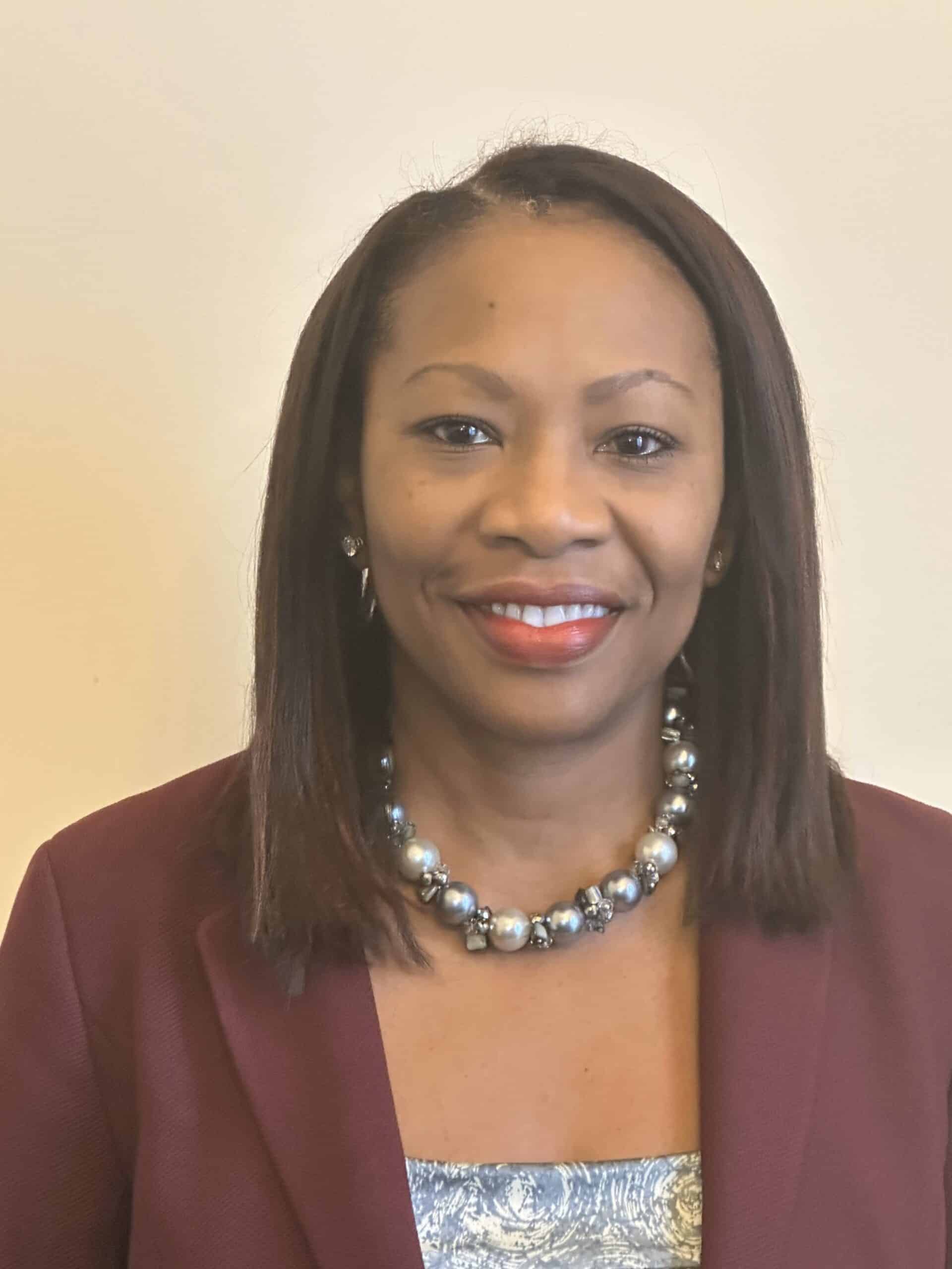 A woman with straight, shoulder-length hair smiles at the camera. She is wearing a maroon blazer, a patterned top, a pearl necklace, and stud earrings, standing against a plain light background.