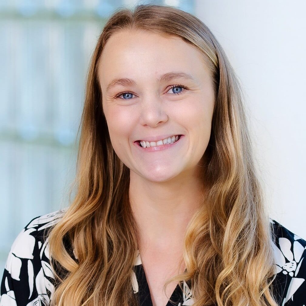 A woman with long, wavy blonde hair and blue eyes smiles at the camera. She is wearing a black blouse with a white leaf pattern. The background is softly blurred with natural light.