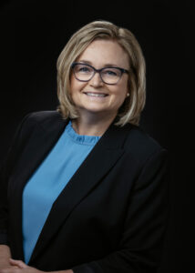 A woman with blonde hair and glasses smiles at the camera. She is wearing a black blazer over a blue blouse and stands against a dark background.