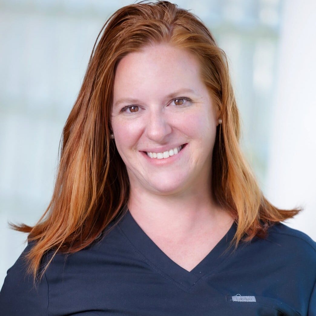 A woman with straight, shoulder-length red hair wearing a dark scrub top smiles at the camera. The background is softly blurred with a light, indoor setting.