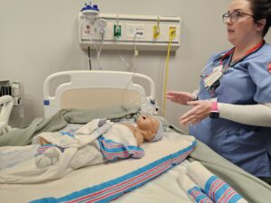 A healthcare worker in scrubs stands beside a hospital bed, explaining or demonstrating care with a swaddled baby mannequin lying on the bed, surrounded by medical equipment.