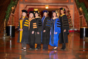 Six women in academic regalia stand in a row indoors, smiling at the camera. The setting is ornately decorated with holiday lights and a Christmas tree in the background.