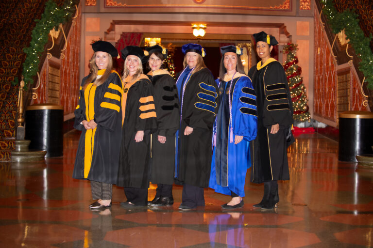 Six women in academic regalia stand in a row indoors, smiling at the camera. The setting is ornately decorated with holiday lights and a Christmas tree in the background.
