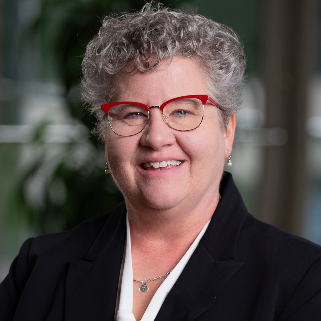 A smiling woman with short curly gray hair, wearing red glasses, a black blazer, and a white blouse, poses in front of a blurred background with green foliage and windows.