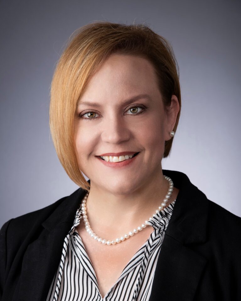 A woman with short, light brown hair smiles at the camera. She is wearing a black blazer, a black-and-white striped blouse, a pearl necklace, and pearl earrings. The background is plain and grey.