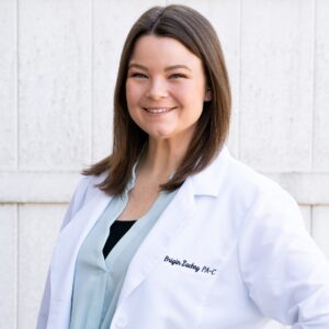 A woman with straight brown hair wearing a white lab coat and light blue blouse stands smiling with one hand on her hip against a white outdoor wall.