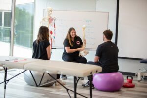 Three people in a classroom sit on exercise balls around a table. One holds a spine model, explaining anatomy. A skeleton and a whiteboard with labeled bones are in the background.