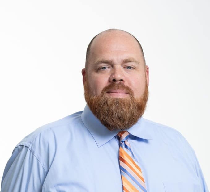 A man with a bald head and a full reddish beard is wearing a light blue dress shirt and a striped orange and blue tie, posing against a plain white background.