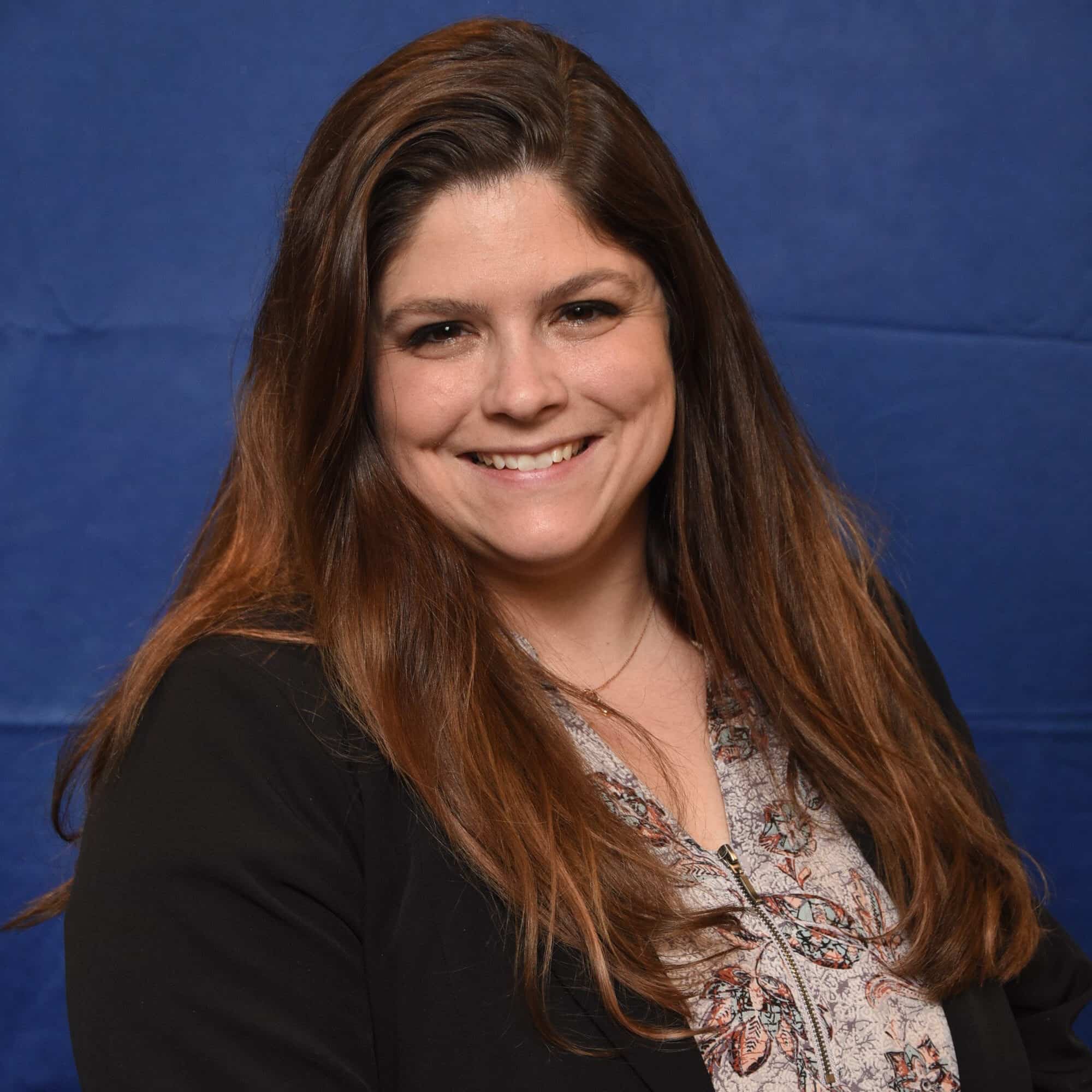 A woman with long brown hair, wearing a black blazer over a floral blouse, smiles while standing in front of a solid blue background.