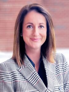A woman with straight, shoulder-length brown hair smiles at the camera. She is wearing a plaid blazer over a dark top, with a blurred brick wall in the background.
