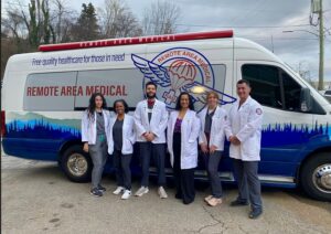 Five healthcare professionals in white coats stand smiling in front of a van labeled "Remote Area Medical," with blue forest graphics and the slogan "Free quality healthcare for those in need" displayed on the roof.