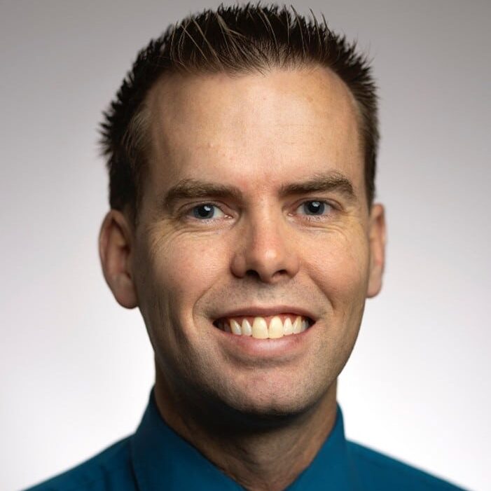 A man with short brown hair smiles at the camera. He is wearing a teal dress shirt and a black tie, with a neutral gray background.