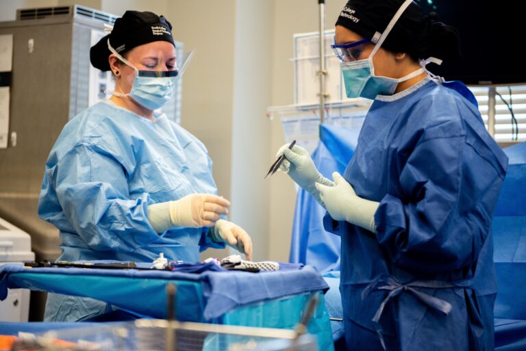 Two surgical technologists wearing blue scrubs, masks, and gloves prepare surgical instruments on a sterile table in an operating room. One hands a tool to the other, both focused on their work.