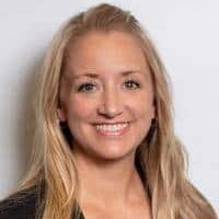 A woman with long blonde hair, wearing a dark blazer over a gray top, smiles at the camera in front of a plain white background.