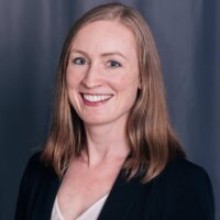 A woman with straight, shoulder-length light brown hair, wearing a black blazer over a light-colored top, smiling in front of a plain dark gray background.