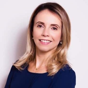 A woman with shoulder-length blonde hair, wearing a navy blue blouse and a gold necklace, smiles at the camera against a plain light background.
