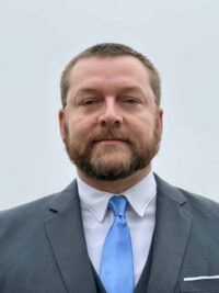 A man with short hair and a beard wearing a grey suit, white shirt, and light blue tie stands against a plain, light-colored background, looking directly at the camera with a neutral expression.