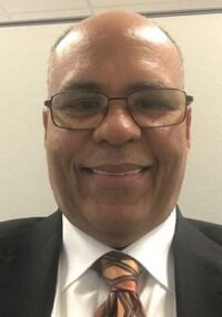 A man wearing glasses, a black suit, white shirt, and a patterned tie smiles at the camera in an indoor setting with a neutral background.