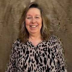 A woman with wavy, shoulder-length hair is smiling. She is wearing a light-colored top with a black, leopard-print pattern, standing in front of a textured brown background.