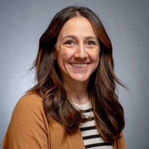 A woman with long brown hair wearing a brown cardigan over a black-and-white striped shirt smiles at the camera against a plain gray background.
