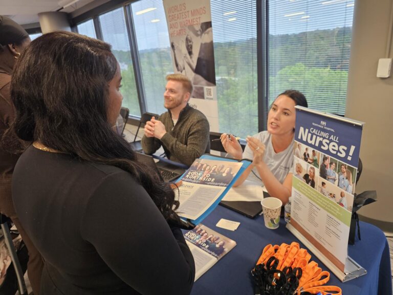 A woman speaks to two recruiters at a table with a “Calling All Nurses!” sign, brochures, and orange lanyards, at a career fair near large windows.