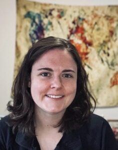 A woman with shoulder-length brown hair smiles at the camera. She is indoors, with a colorful abstract tapestry hanging on the wall behind her.