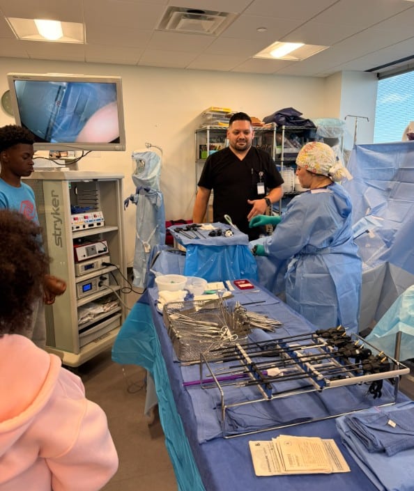Medical staff in scrubs and surgical masks stand beside a table with surgical instruments, conducting a demonstration. A monitor displays an internal view. Two observers watch in the foreground. The room is well-lit with large windows.