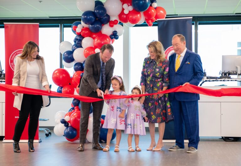 Five adults and two young girls cut a large red ribbon at an indoor ceremony, with red, white, and blue balloons in the background. The girls wear matching dresses and hold the scissors together.