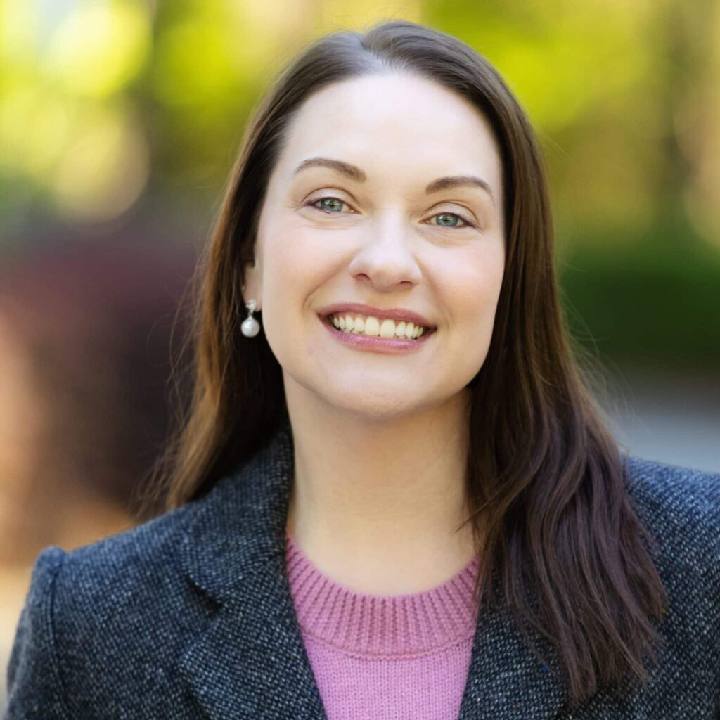 A woman with straight brown hair, wearing a gray blazer over a pink sweater, smiles at the camera outdoors with a blurred green and brown background.